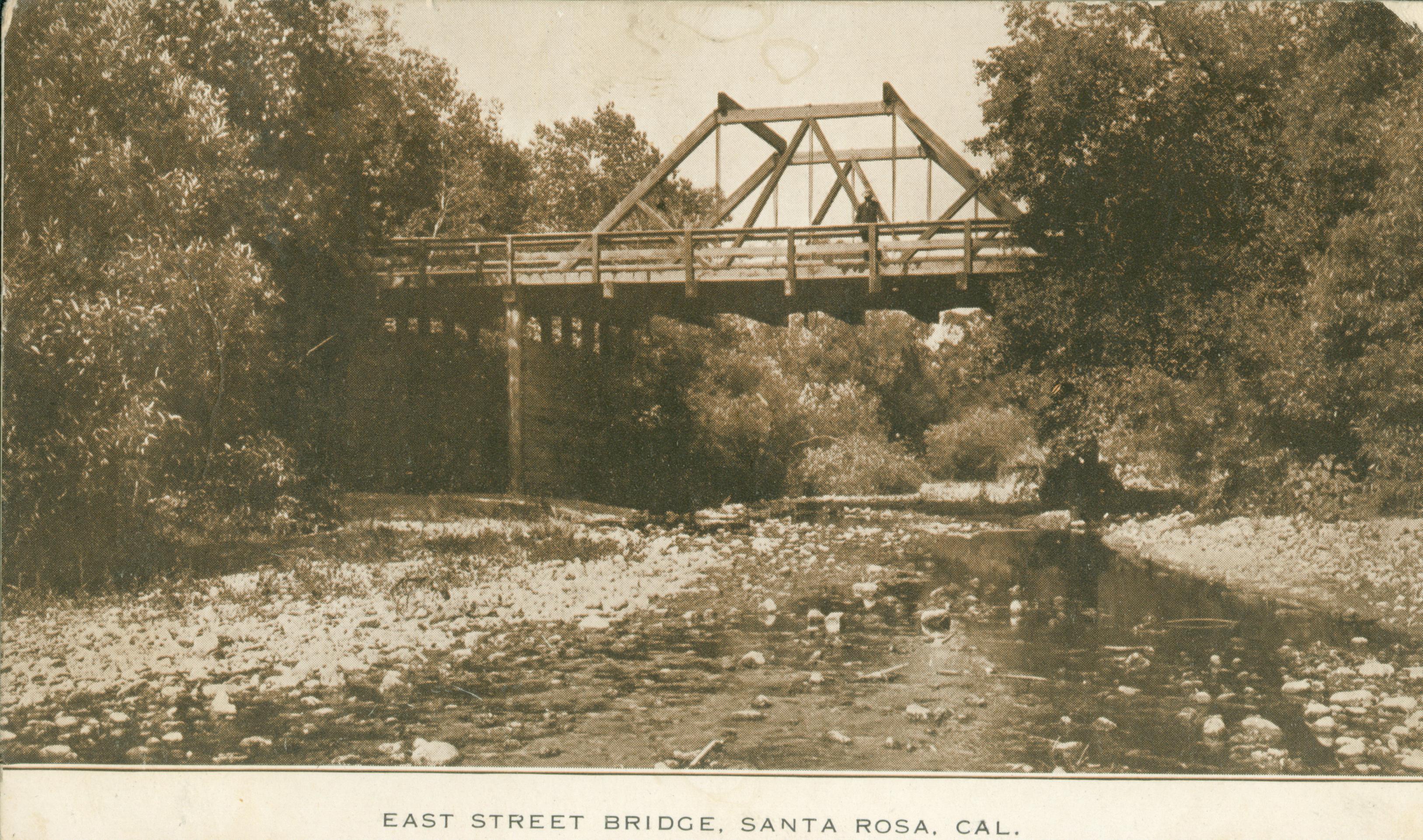 Shows a street bridge spanning a river lined by trees